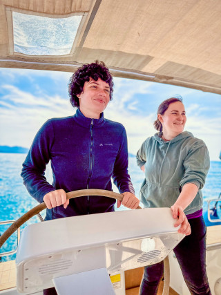 Zwei Menschen, die auf einem Segelboot stehen und lächeln. Eine Person hat das Schiffsruder in der Hand.